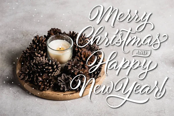 Scented candle with pine cones on wooden plate near merry christmas and happy new year lettering on grey background — Stock Photo