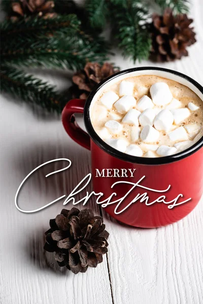 Red cup of cocoa near pine cone and merry christmas lettering on blurred wooden background — Stock Photo
