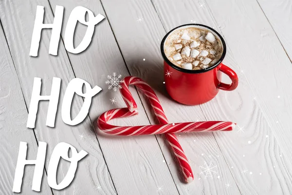 Tazza rossa di cacao vicino bastoncini di zucchero natalizio vicino ho ho ho ho lettering su sfondo di legno — Foto stock