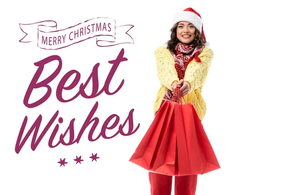 Joyful young woman in santa hat and scarf with ornament holding red shopping bags near merry christmas, best wishes lettering on white — Stock Photo