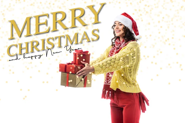 Joyful young woman in santa hat, scarf and knitted sweater holding gifts near merry christmas and happy new year lettering on white — Stock Photo