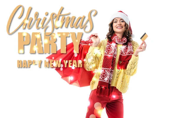 Fröhliche Frau in Weihnachtsmütze und Schal mit Ornament, die Einkaufstaschen und Kreditkarte in der Nähe der Weihnachtsfeier hält, frohes neues Jahr auf weiß — Stockfoto