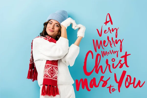 Jovem mulher satisfeita em cachecol quente, luvas e chapéu mostrando sinal de coração perto de um Natal muito alegre para você lettering em azul — Fotografia de Stock