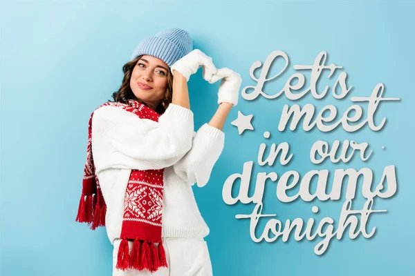 Pleased young woman in warm scarf, gloves and hat showing heart sign near lets meet in our dreams tonight lettering on blue — Stock Photo