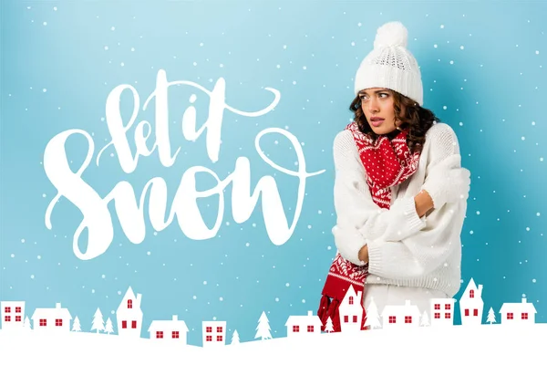 Freezing young woman in winter outfit embracing herself near let it snow lettering and houses illustration on blue — Stock Photo
