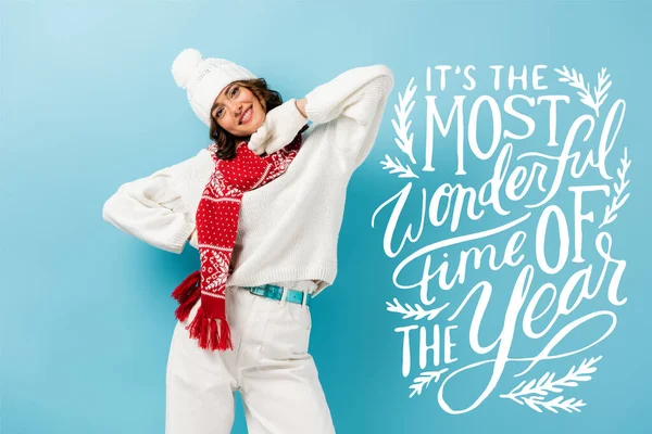 Pleased young woman in white winter outfit, warm scarf, gloves and hat standing near its the most wonderful time of the year lettering on blue — Stock Photo