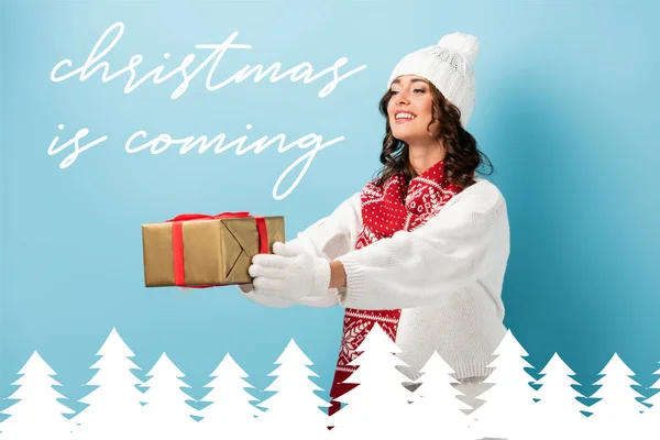 Jeune femme heureuse en tenue d'hiver avec les mains tendues tenant présent enveloppé près de Noël est à venir lettrage sur bleu — Photo de stock