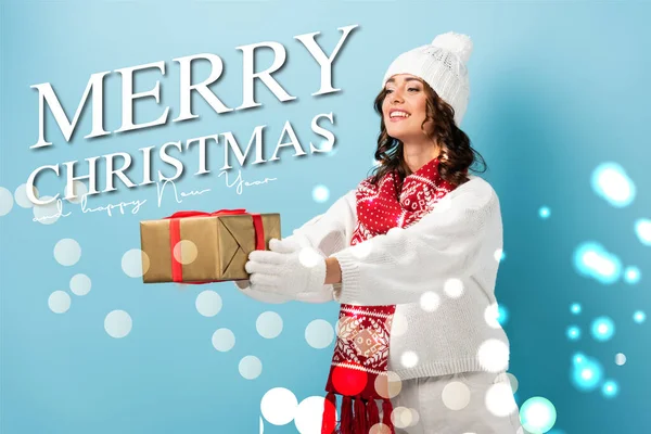 Young pleased woman in winter outfit with outstretched hands holding wrapped present near merry christmas and happy new year lettering on blue — Stock Photo