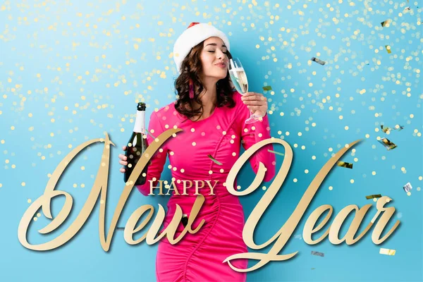 Young woman in santa hat holding bottle and drinking champagne near confetti and happy new year lettering on blue — Stock Photo