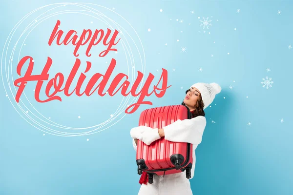 Young woman in winter outfit carrying heavy suitcase near happy holidays lettering on blue — Stock Photo
