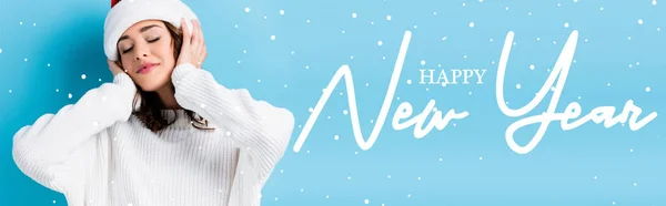 Young brunette woman with closed eyes adjusting hat near happy new year lettering on blue, banner — Stock Photo