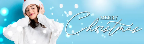 Young brunette woman with closed eyes adjusting hat near merry christmas lettering on blue, banner — Stock Photo