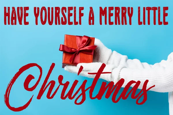 Cropped view of young woman in gloves holding gift box near have yourself a merry little christmas lettering on blue — Stock Photo