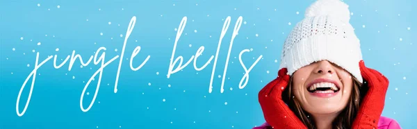 Joyful woman in red gloves covering eyes while adjusting white hat near jingle bells lettering on blue, banner — Stock Photo