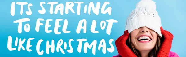 Joyful woman in red gloves covering eyes while adjusting white hat near its starting to feel a lot like christmas lettering on blue, banner — Stock Photo