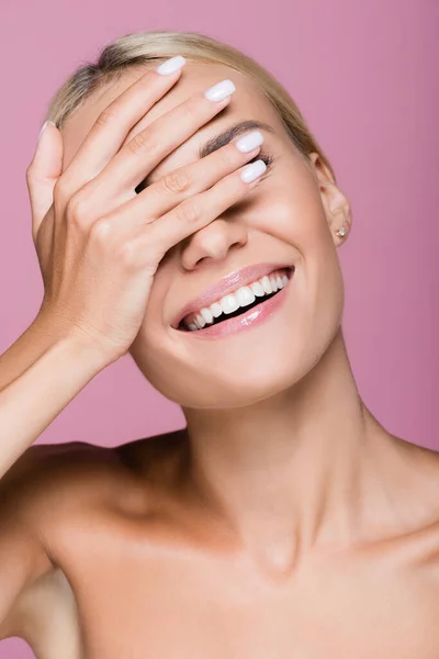 Усміхнена красива блондинка позує з рукою на обличчі ізольована на рожевому — стокове фото