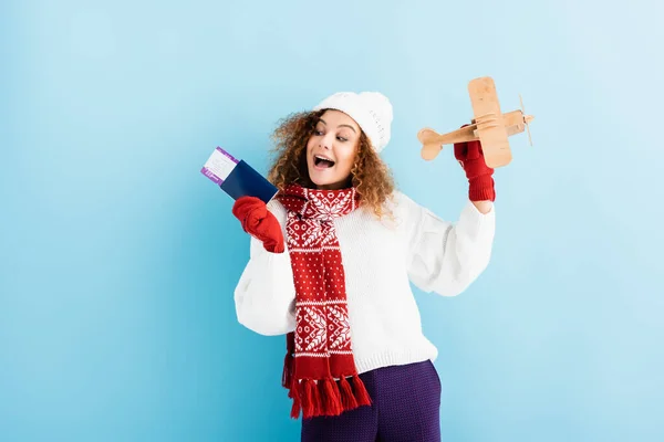 Excited young woman in hat and sweater holding passport and toy plane on blue — Stock Photo