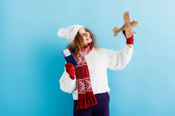 Smiling young woman in hat and sweater holding passport and toy plane on blue — Stock Photo