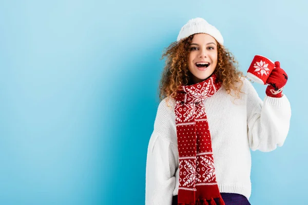 Excité jeune femme en chapeau et pull tenant tasse en tricot titulaire sur bleu — Photo de stock