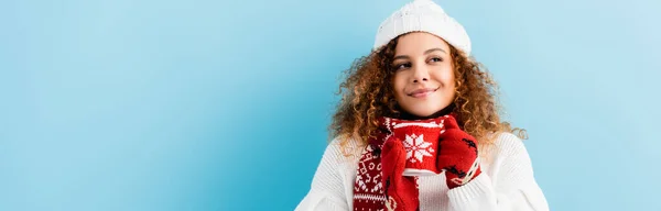 Mujer alegre y rizada en sombrero y suéter sosteniendo taza de té en titular de punto en azul, bandera - foto de stock