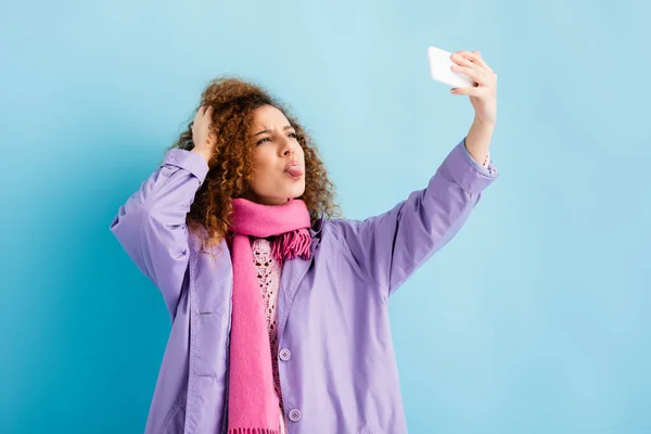 Кудрявая молодая женщина в зимнем пальто и розовый вязаный шарф держа смартфон и торчащий язык, делая селфи на синий — стоковое фото