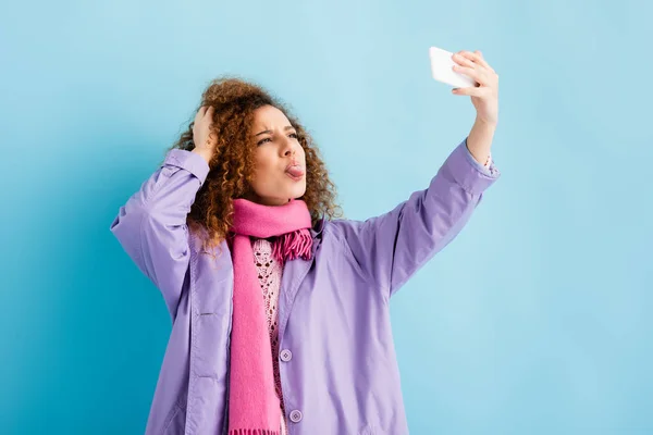 Mujer joven y rizada con abrigo de invierno y bufanda de punto rosa sosteniendo el teléfono inteligente y sacando la lengua mientras toma selfie en azul — Stock Photo
