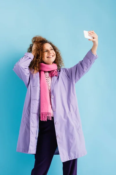 Cheerful young woman in winter coat and pink knitted scarf holding smartphone and taking selfie on blue — Stock Photo