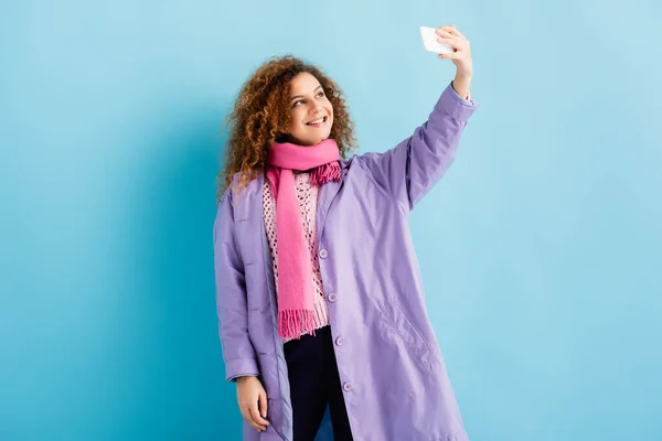 Mujer joven feliz en abrigo de invierno y bufanda de punto rosa sosteniendo teléfono inteligente y tomando selfie en azul - foto de stock