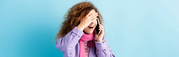 Scioccato giovane donna in cappotto invernale e rosa sciarpa lavorata a maglia parlando su smartphone su blu, banner — Foto stock
