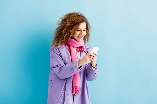 Happy young woman in winter coat and pink knitted scarf texting on smartphone on blue — Stock Photo
