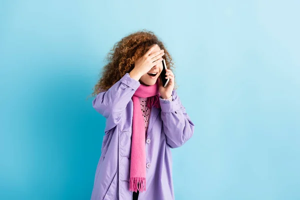 Schockierte junge Frau in Wintermantel und rosa Strickschal redet auf Smartphone auf blau — Stockfoto