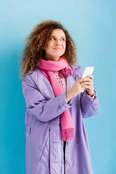 Mujer joven pensativa en abrigo de invierno y rosa bufanda de punto con teléfono inteligente en azul - foto de stock