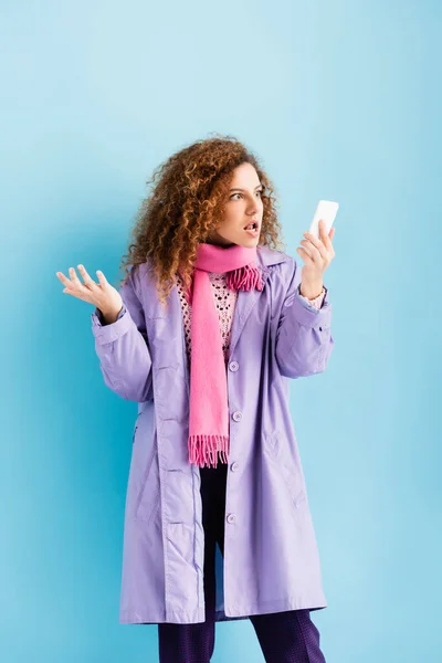 Сердитая молодая женщина в зимнем пальто и розовый вязаный шарф глядя на смартфон на синий — стоковое фото