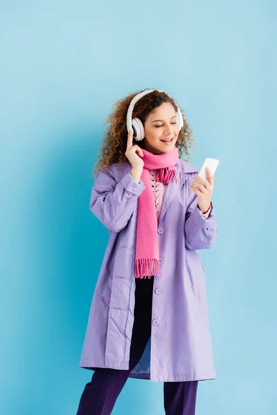 Jovem feliz em fones de ouvido sem fio, casaco de inverno e lenço de malha rosa usando smartphone em azul — Fotografia de Stock