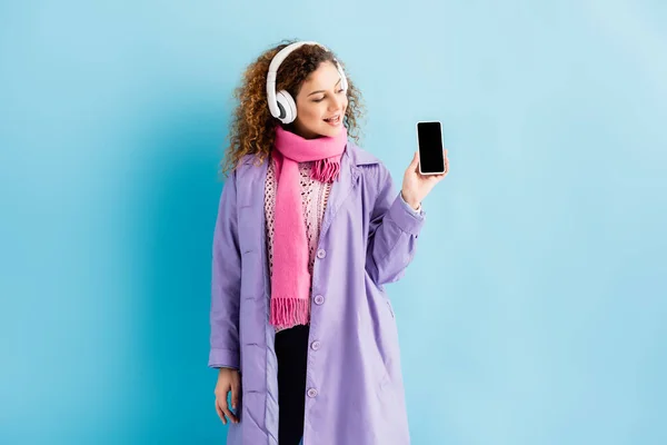 Щаслива молода жінка в бездротових навушниках, зимове пальто і рожевий в'язаний шарф дивиться на смартфон з порожнім екраном на синьому — стокове фото