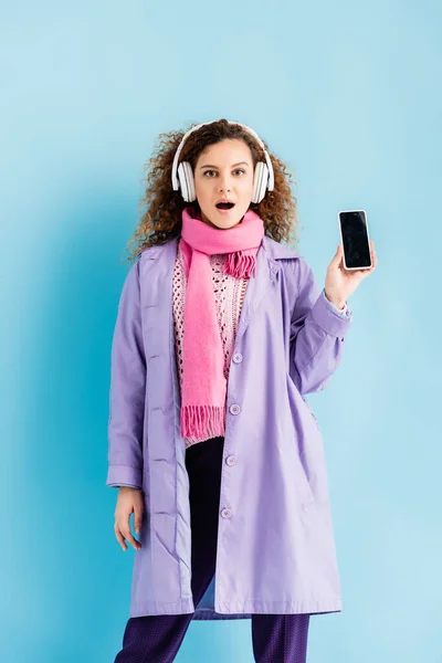 Chocado jovem mulher em fones de ouvido sem fio, casaco de inverno e rosa de malha cachecol segurando smartphone com tela em branco no azul — Fotografia de Stock