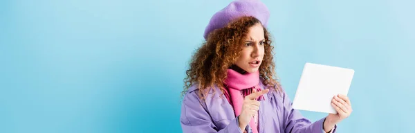 Unzufriedene junge Frau in Wintermantel, Baskenmütze und rosafarbenem Strickschal zeigt mit dem Finger auf digitales Tablet auf blau, Banner — Stockfoto