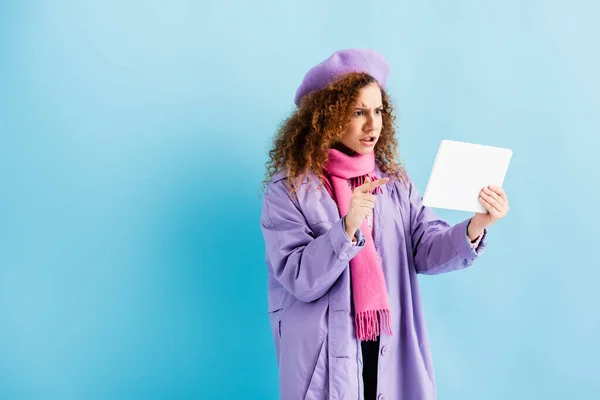 Giovane donna insoddisfatta in cappotto invernale, berretto e sciarpa a maglia rosa che punta con il dito al tablet digitale su blu — Foto stock