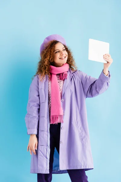 Allegra giovane donna in cappotto invernale, berretto e rosa sciarpa lavorata a maglia con tablet digitale e scattare selfie su blu — Foto stock