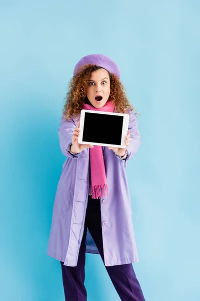 Shocked young woman in winter coat, beret and pink knitted scarf holding digital tablet with blank screen on blue — Stock Photo