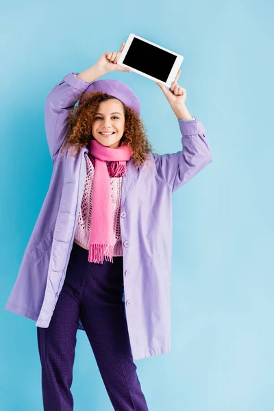 Happy young woman in winter coat, beret and pink knitted scarf holding digital tablet with blank screen above head on blue — Stock Photo