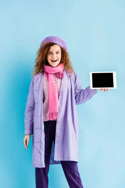Aufgeregte Frau in Wintermantel, Baskenmütze und rosa Strickschal mit digitalem Tablet und leerem Bildschirm auf blauem Hintergrund — Stockfoto