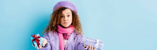 Curly woman in winter coat, beret and pink knitted scarf holding christmas presents on blue, banner — Stock Photo