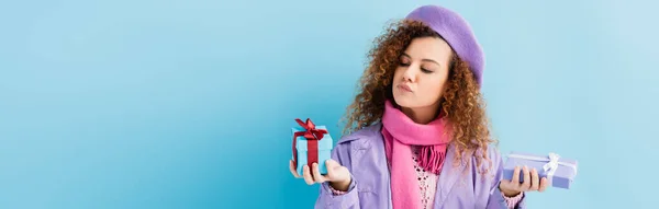 Curly woman in winter coat, beret and pink knitted scarf choosing between christmas presents on blue, banner — Stock Photo