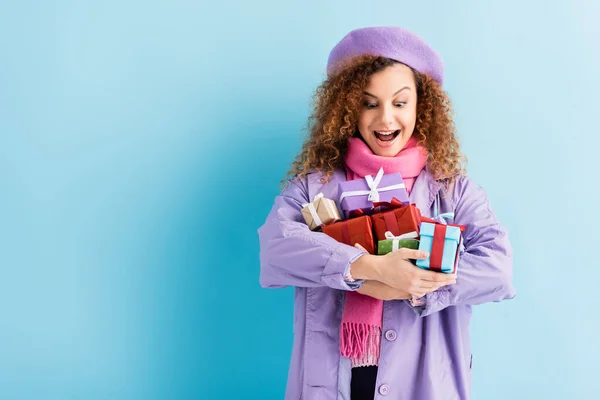 Excited woman in winter coat, beret and pink knitted scarf holding christmas presents on blue — Stock Photo