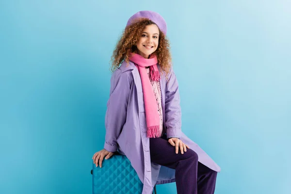 Happy woman in beret, scarf and coat smiling while sitting on suitcase on blue — Stock Photo