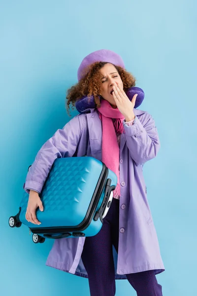 Tired woman in beret, scarf, travel pillow and coat holding suitcase and yawning on blue — Stock Photo