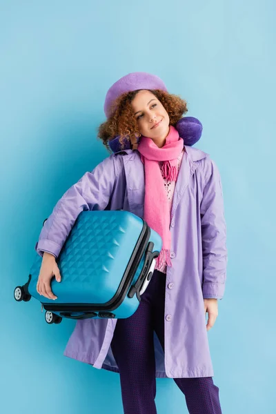 Sleepy woman in beret, scarf, travel pillow and coat holding suitcase on blue — Stock Photo