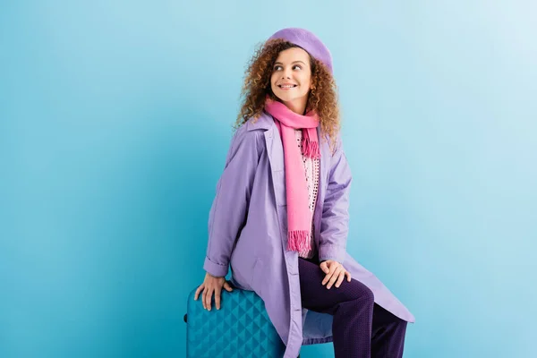 Весела жінка в береті, шарфі, подушці для подорожей і пальто, сидячи на багажі на синьому — стокове фото