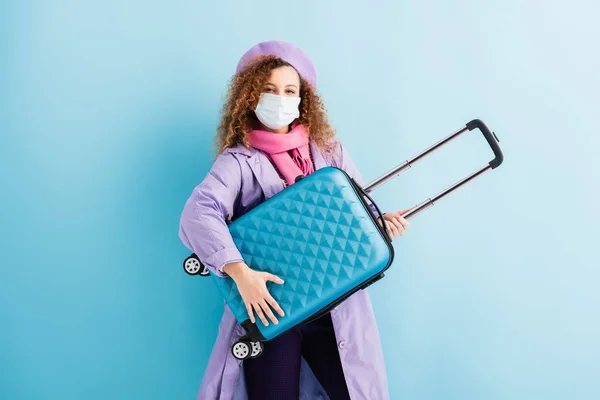 Mujer rizada en boina, bufanda, máscara médica y abrigo que sostiene la maleta en azul - foto de stock
